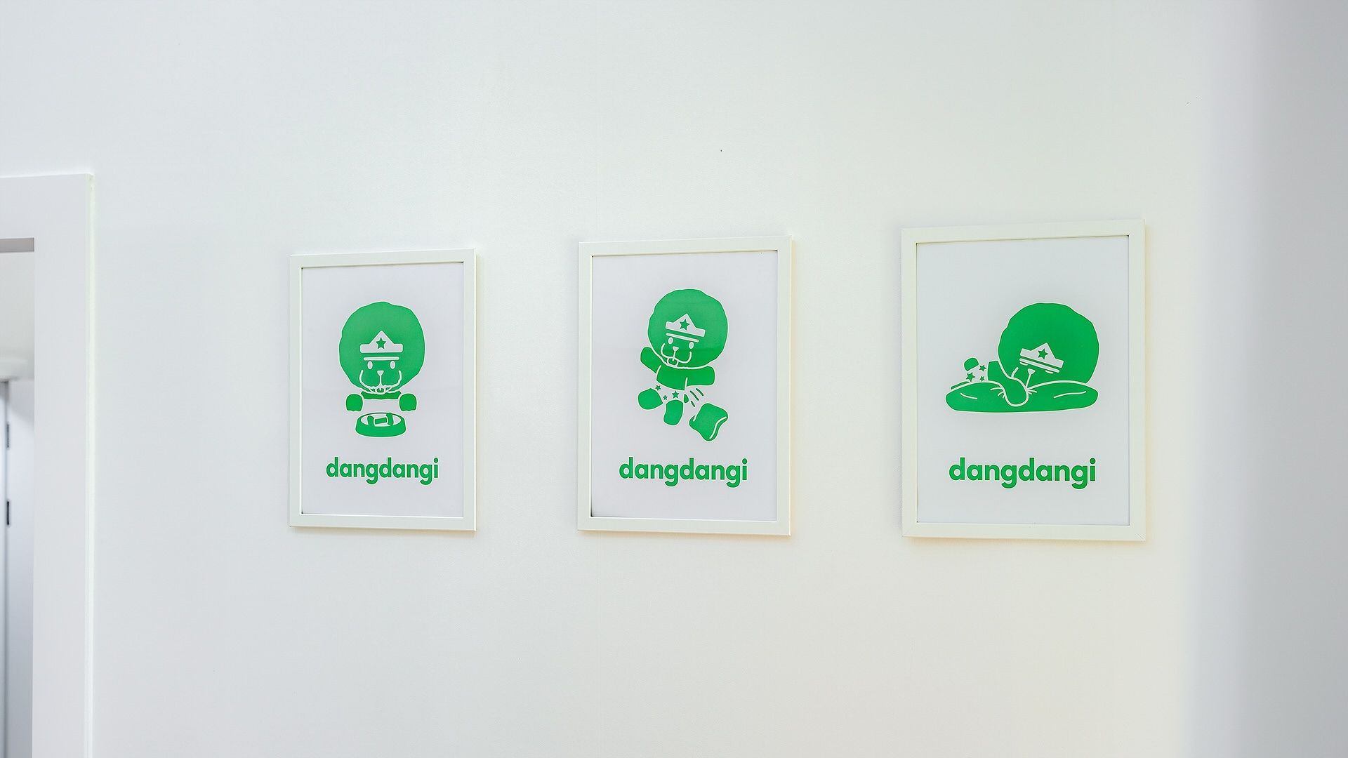 Trois œuvres d’art encadrées, représentant des figures vertes, et sur lesquelles on peut lire le mot « dangdangi », le tout accroché à un mur blanc.