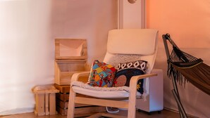 Een gezellige kamer met een witte fauteuil, kleurrijke kussens, een houten bijzettafeltje en een hangstoel.