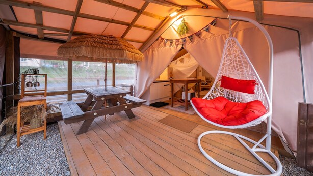 Een gezellig interieur met een houten terras, een hangstoel met rode kussens, een rieten dak en een picknicktafel.