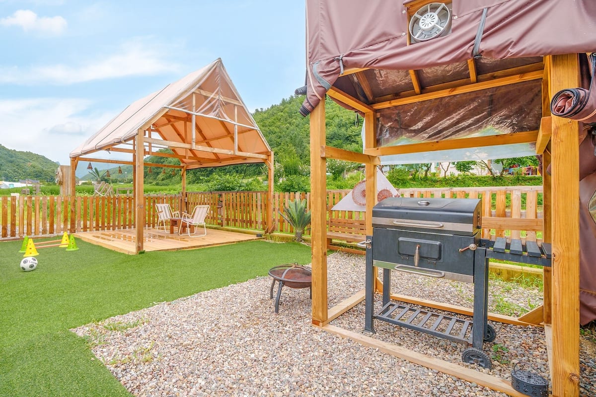 Una terraza de madera con una parrilla, un espacio de estar techado y un pequeño campo de fútbol.