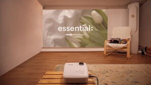 Ein Raum mit einem Projektor auf einem Holztisch, einem Stuhl mit farbenfroher Sitzkissen und einer Wand mit großem Blumenmuster.