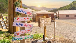 Un panneau indicateur en bois, doté de plusieurs flèches indiquant différentes directions, dont les inscriptions « Bienvenue à Illoo » et « Zone verte », le tout sur fond de tentes et d’un paysage montagneux.