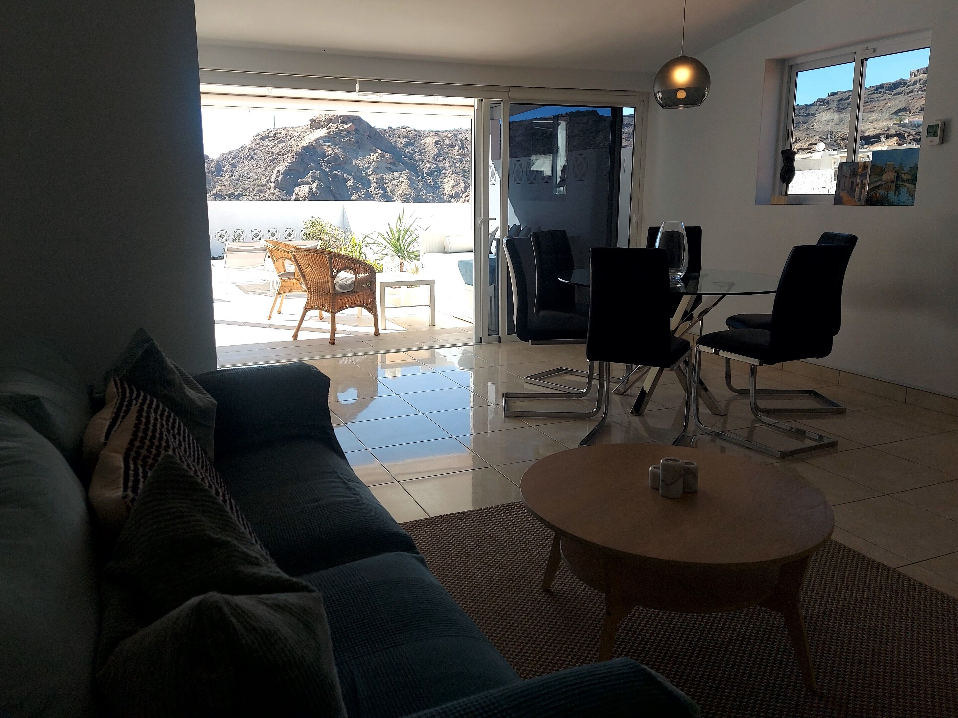 Ein Wohnzimmer mit einer Couch, einem Couchtisch und einem Essbereich mit Stühlen und einem Tisch. Durch ein großes Fenster ist eine Bergblick zu sehen.