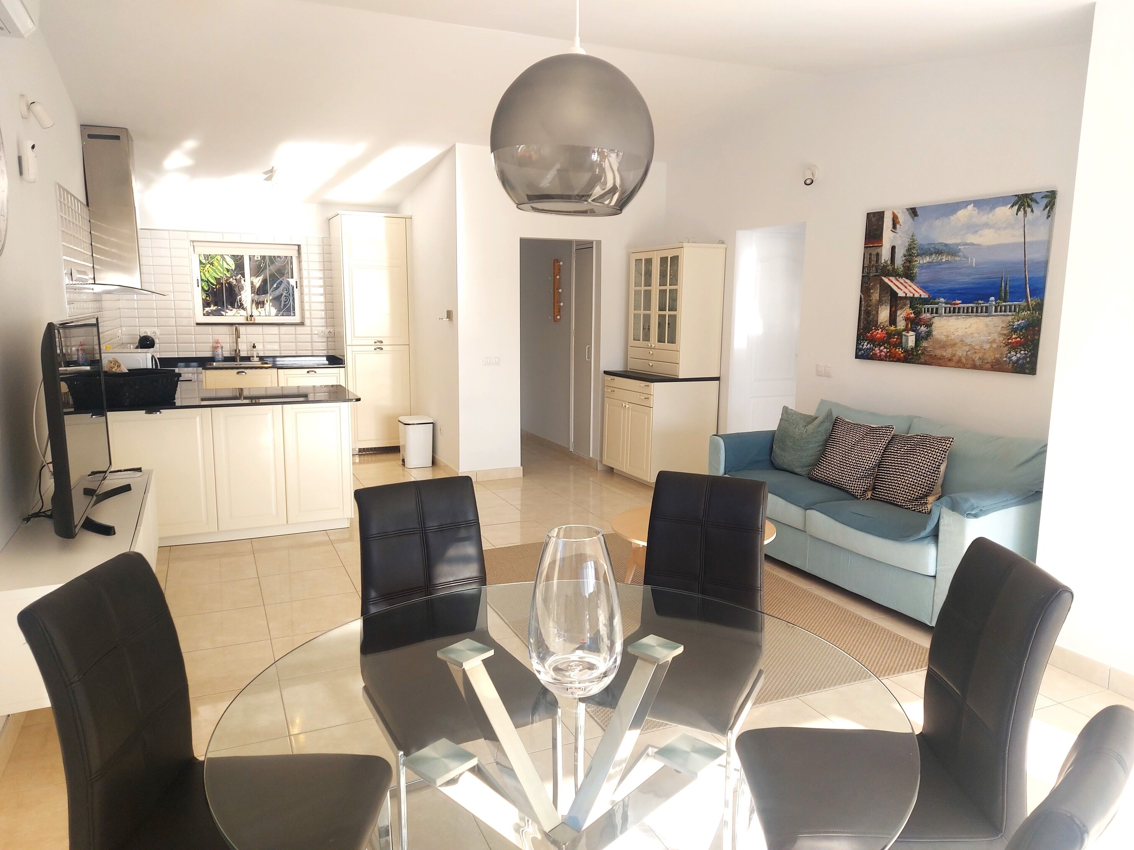 Een moderne woonruimte met een eetgedeelte, bestaande uit een glazen tafel en zwarte stoelen, een lichtblauwe bank met opvallende kussens, en een keuken met witte kastjes.