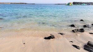 Ein Strand mit klarem Wasser, Felsen und einer in der Ferne liegenden Felsformation.