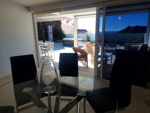 Une pièce moderne avec une table et des chaises en verre, un verre à vin, et une vue sur l’océan et une montagne.