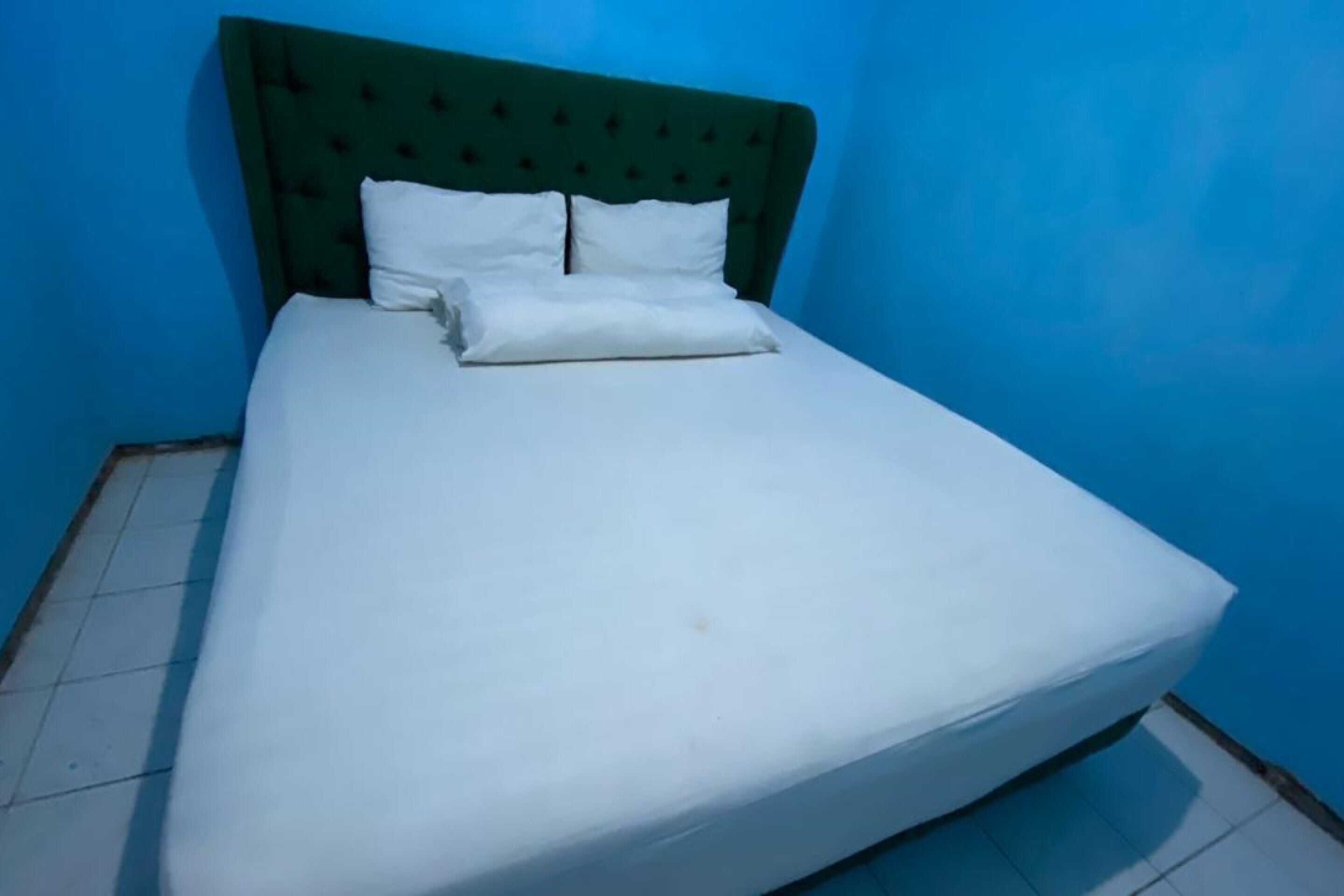 Een eenpersoonsbed met een groen gestoffeerd hoofdeinde en witte beddengoed in een kamer met blauwe wanden en betegelde vloer.
