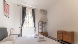 A neatly arranged hotel room with a bed, a bedside table with a lamp, a wooden dresser, and a window with curtains.