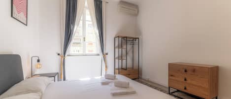 A neatly arranged hotel room with a bed, a bedside table with a lamp, a wooden dresser, and a window with curtains.