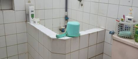A small bathroom with white tiles, a toilet, a sink, and a shower area. There are toiletries on the sink and a sign on the wall.