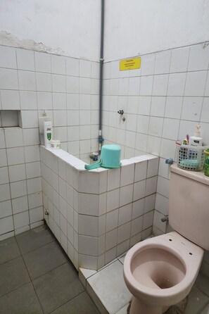 A small bathroom with white tiles, a toilet, a sink, and a shower area. There are toiletries on the sink and a sign on the wall. - Hotel O Regina Homestay Syariah (Surabaya)