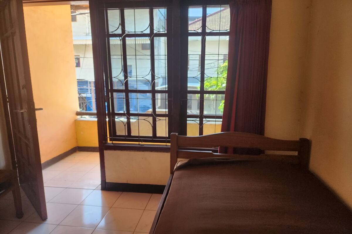 Ein Zimmer mit einem Bett, einem Fenster mit Gitterstäben und Blick auf ein Gebäude.