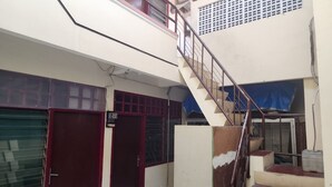 A narrow corridor with a staircase leading to an upper level. There are two doors with red frames, one of which is closed. The walls are painted in a light color, and there is a blue tarp covering part of the upper level. - Hotel O Regina Homestay Syariah (Surabaya)