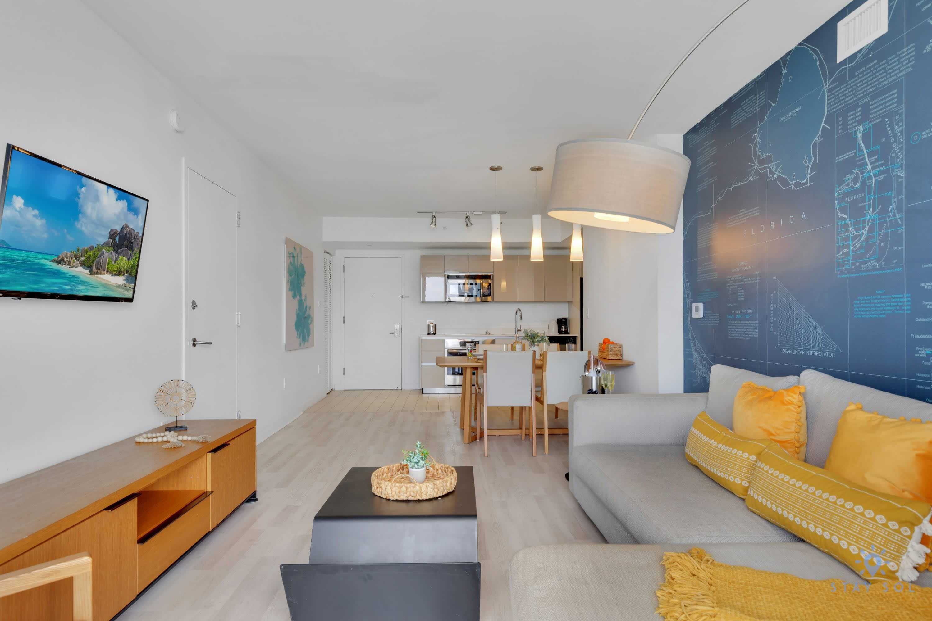 Una sala de estar moderna con sofá, mesa de centro y televisor. Se observa una cocina al fondo con comedor.