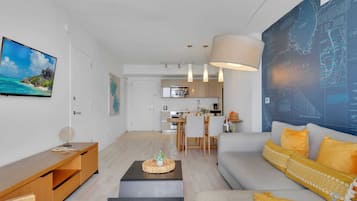 Una sala de estar moderna con sofá, mesa de centro y televisor. Se observa una cocina al fondo con comedor.