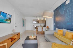 Una sala de estar moderna con sofá, mesa de centro y televisor. Se observa una cocina al fondo con comedor.