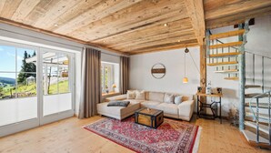 Ein geräumiges Wohnzimmer mit großem Fenster, Holzboden, einer Sofagarnitur, einem roten Teppich mit Muster und einer Treppe.