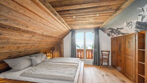 Ein Schlafzimmer mit schrägen Wänden, Holzwänden, einem Bett mit grauer Bettwäsche, einem Holzschrank und Blick durch ein Fenster.