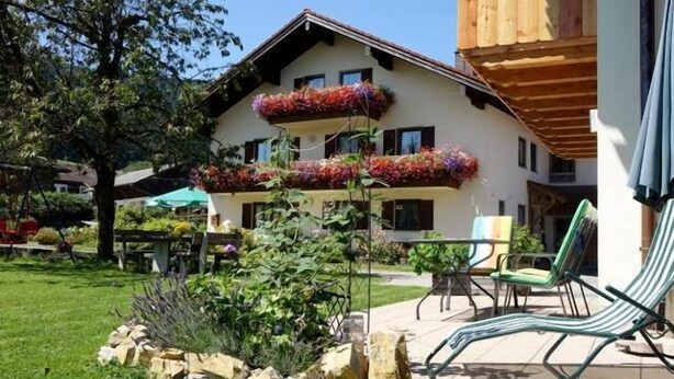 A two-story building with balconies adorned with colorful flowers, a patio area with lounge chairs, and a well-maintained garden. - Ferienwohnung Gaisberg (Ainring)