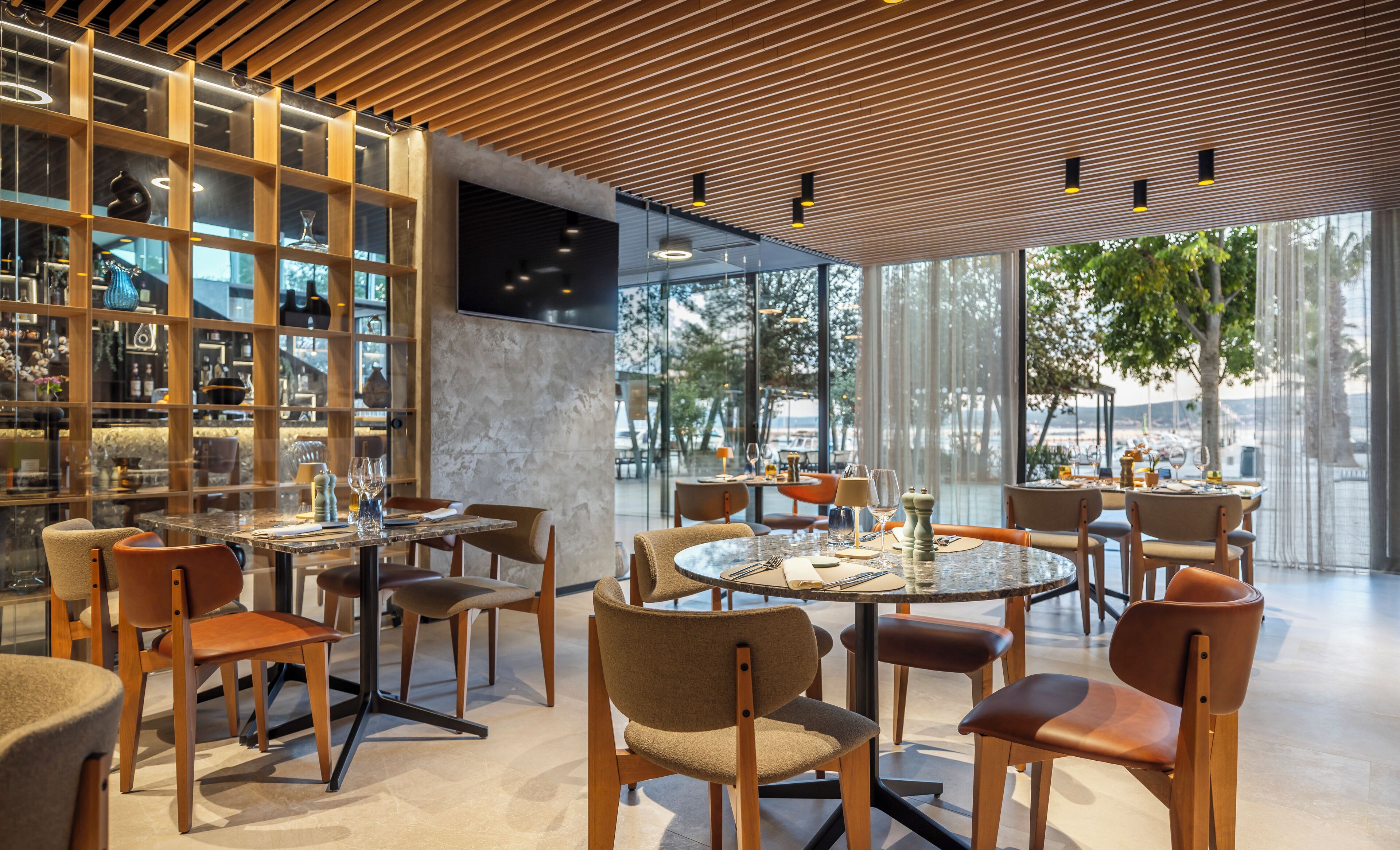 Un intérieur de restaurant moderne avec des tables rondes, des chaises rembourrées et une vue sur la mer grâce à de grandes fenêtres.