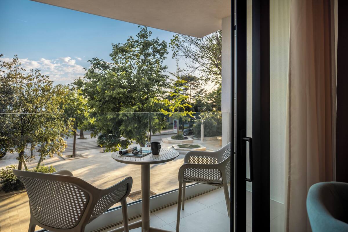A balcony with two chairs, a table, and a view of a street and trees.