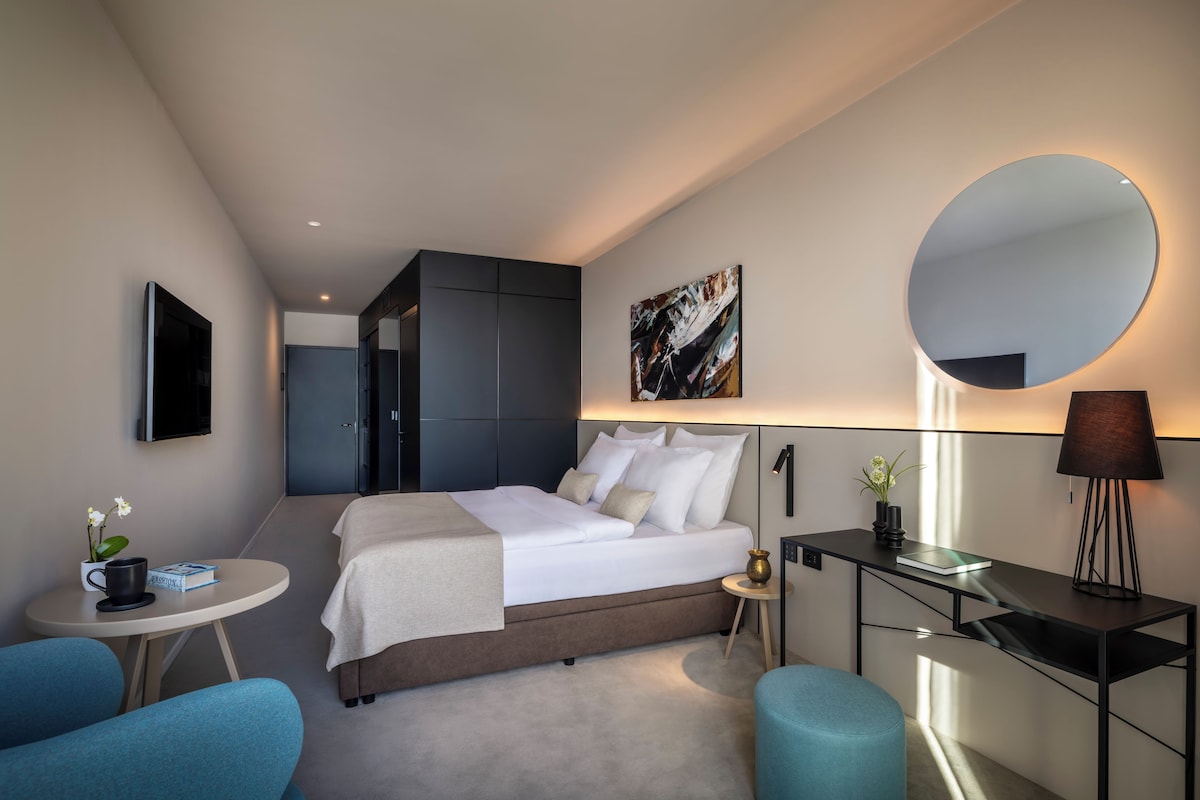 Ein modernes Hotelzimmer mit einem Bett, einem Schreibtisch, einem runden Spiegel und einem kleinen Tisch mit einer Lampe.