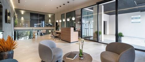 A modern reception area with a wooden desk, two grey armchairs, a round table with a book and flowers, and a large glass wall with a view of the outside.