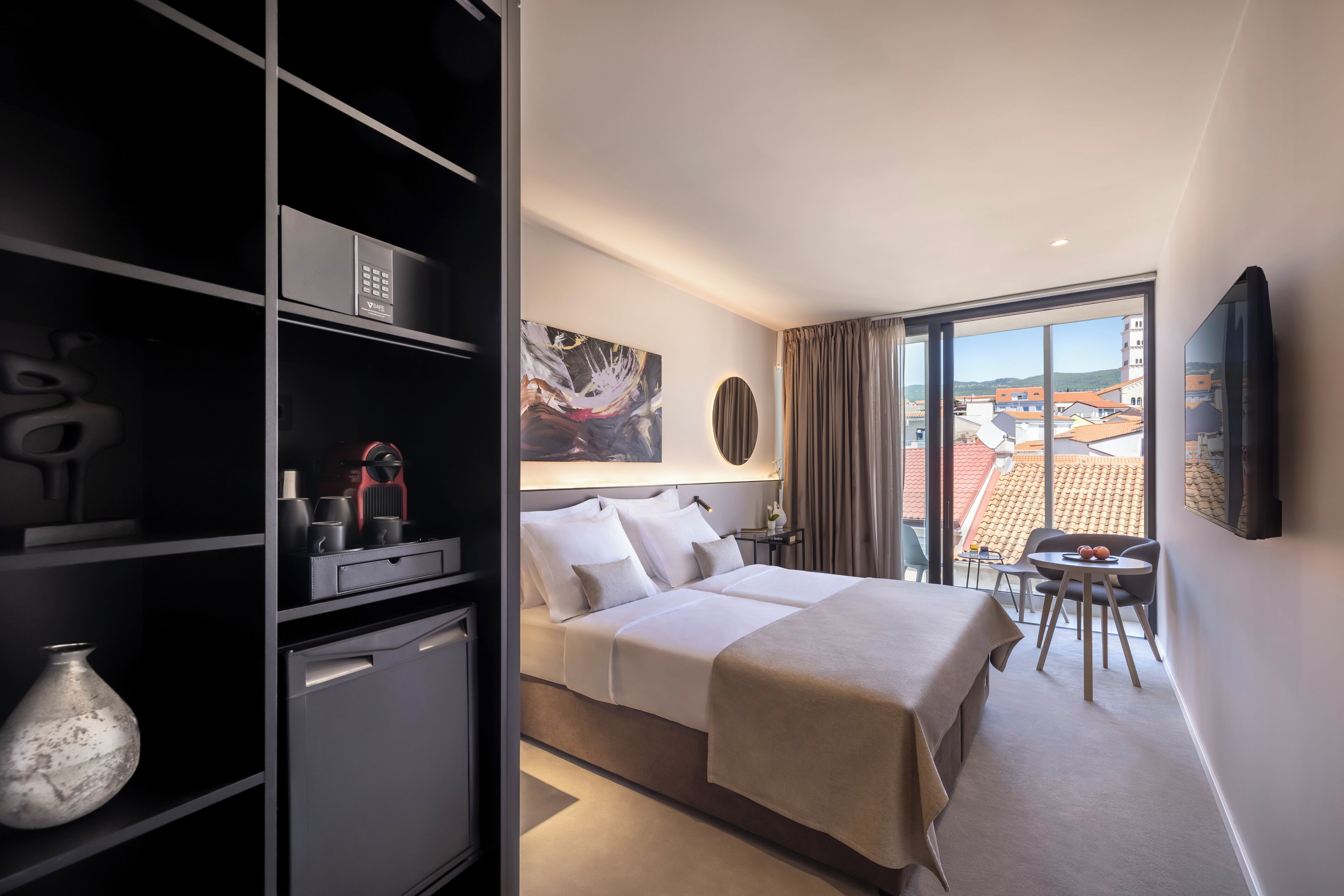 A hotel room with a bed, a desk, a chair, a TV, and a view of buildings.