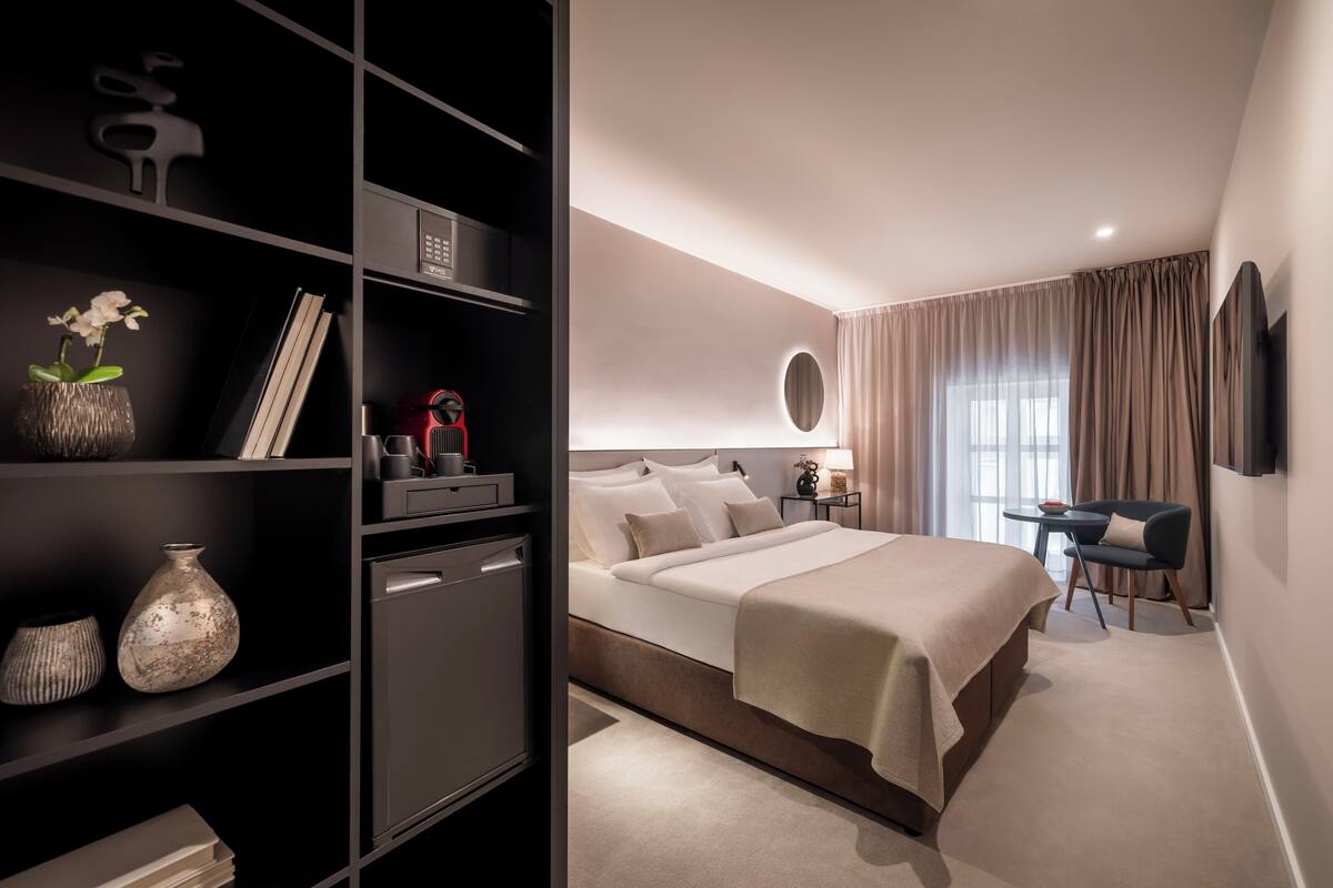 A modern hotel room with a large bed, a desk, a bookshelf, and a TV mounted on the wall.