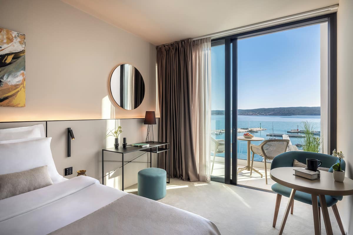 A modern hotel room with a large bed, a desk, a chair, and a view of the sea.