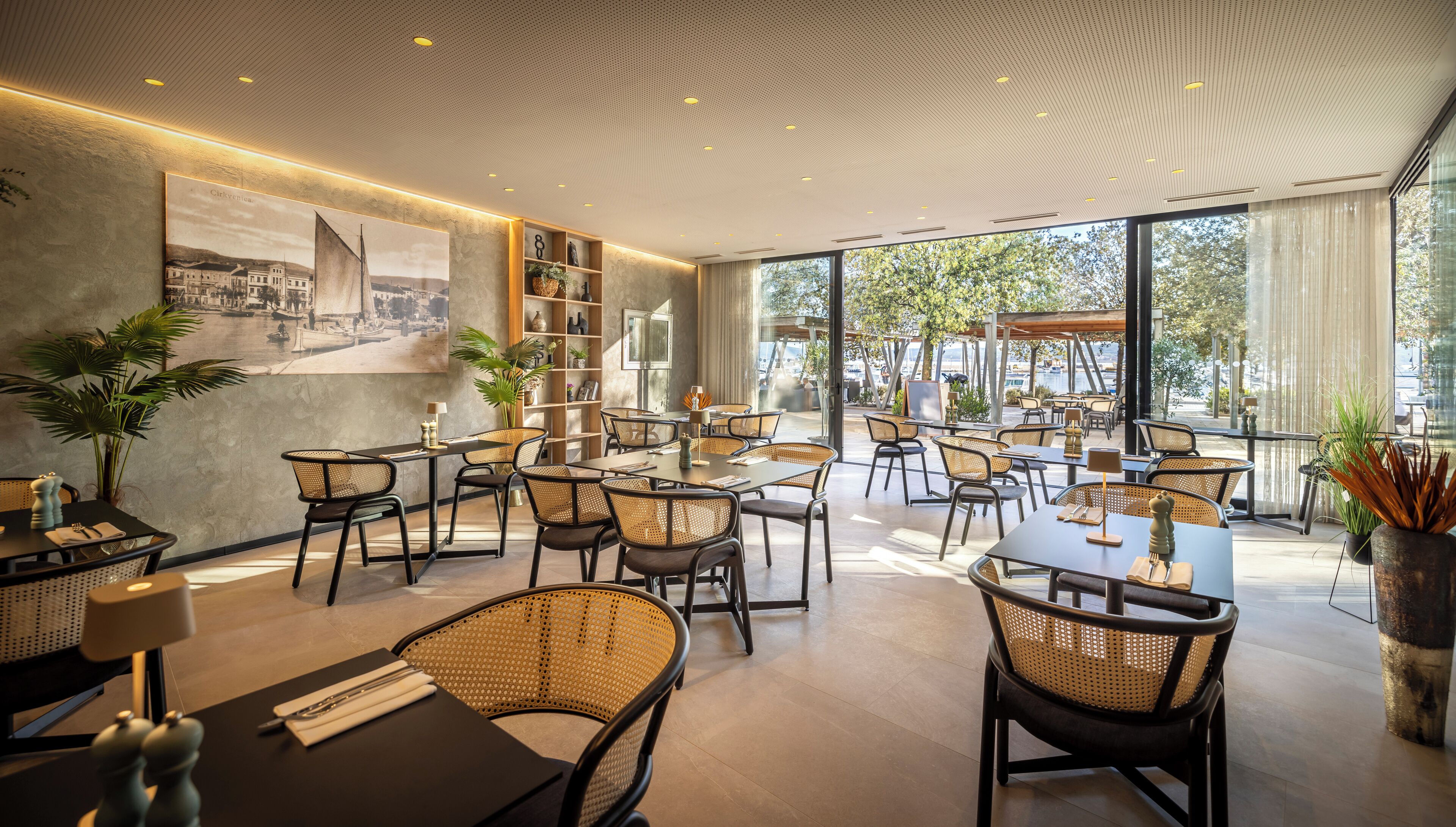 Ein modernes Restaurant mit Korbstühlen, Tischen und einem großen Fenster mit Blick auf ein Gewässer.
