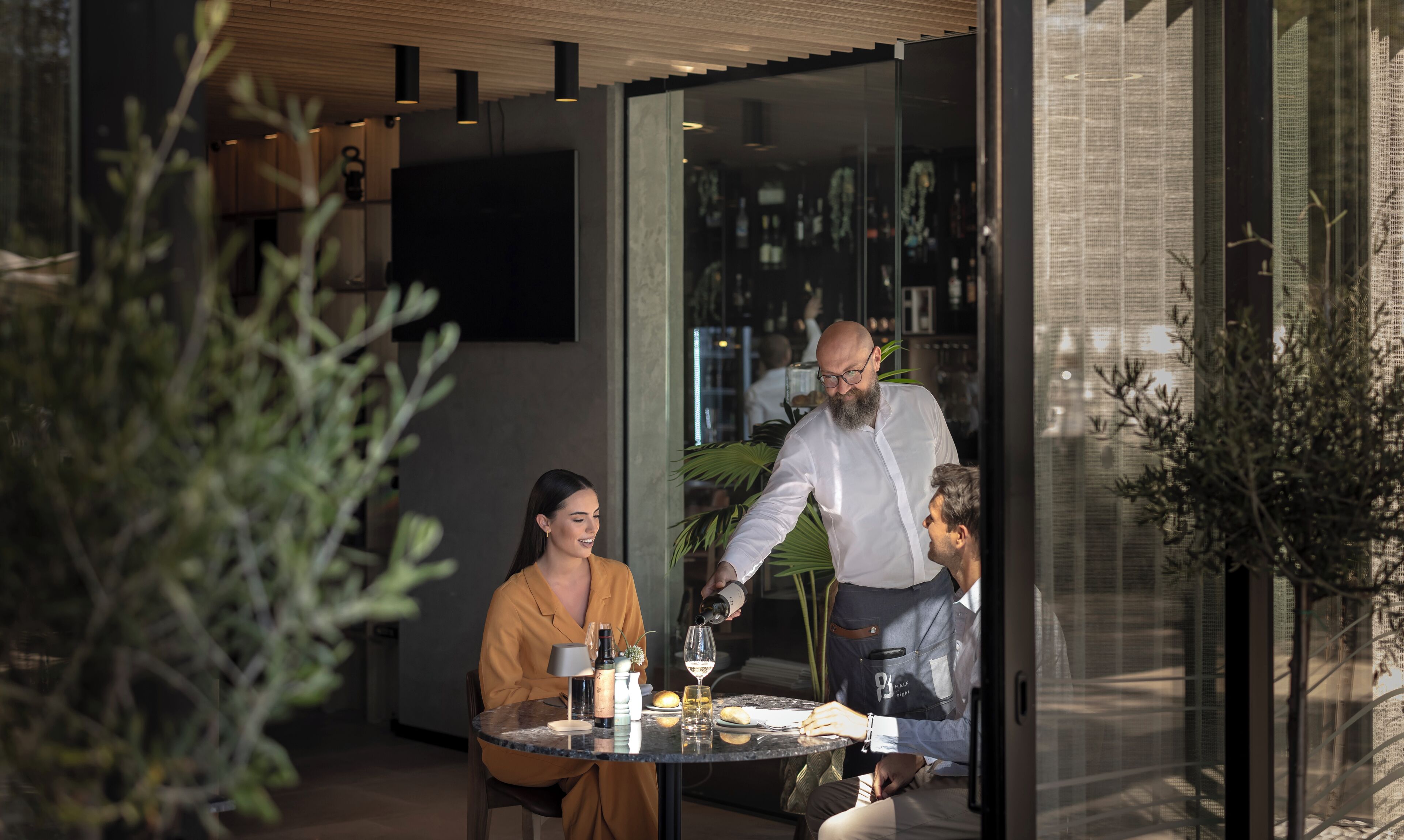 An outdoor dining area with a glass partition, a table with wine glasses, and a person pouring a drink.