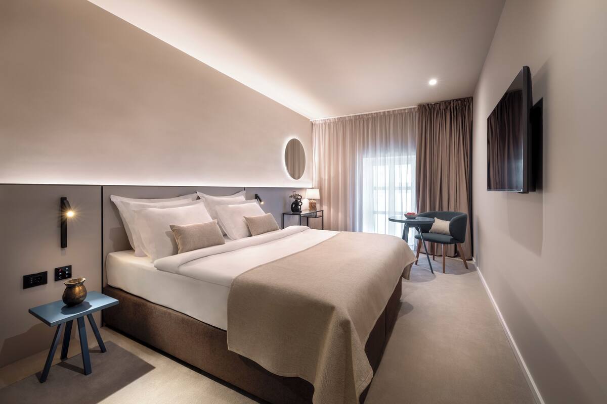 A modern hotel room with a large bed, a small table with a lamp, a TV mounted on the wall, and a view of the outdoors through the window.