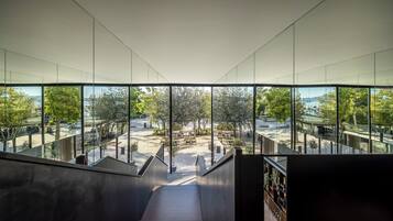 Un interno moderno con una scala che conduce a un'area esterna con tavoli e sedie, ampie finestre e vista su alberi e uno specchio d'acqua.