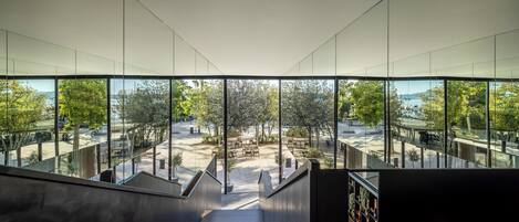 Un interior moderno con una escalera que conduce a un espacio exterior con mesas y sillas, ventanas amplias y vistas a árboles y una masa de agua.