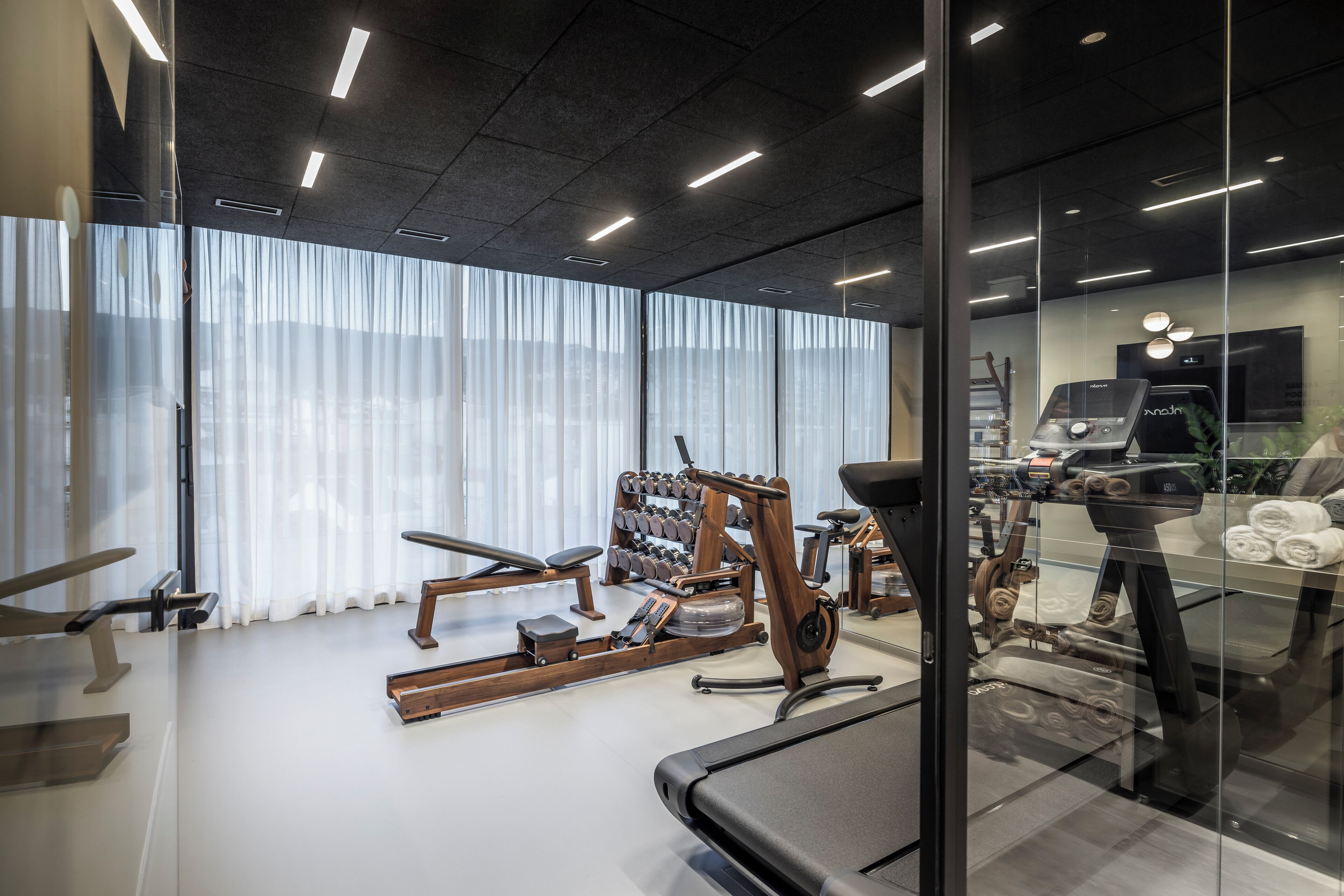 A modern gym with various exercise equipment, large windows with sheer curtains, and a clean, well-lit interior.