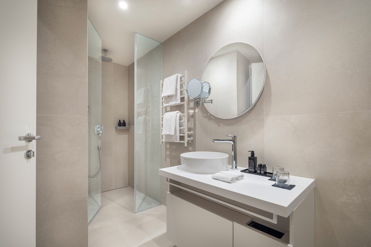 Une salle de bain moderne avec une douche à l’italienne en verre, un meuble-lavabo blanc avec un miroir rond et un grand miroir rond placé au-dessus du lavabo.
