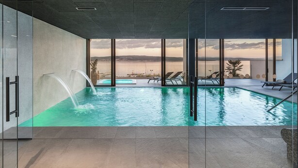 A modern indoor pool with clear glass walls, a view of the sea, and lounge chairs. - Aminess Younique Narrivi Hotel (Crikvenica)