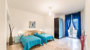 Ein Schlafzimmer mit zwei Betten, einem Nachttisch, einem Wandbild und einem Fenster mit blauen Vorhängen.