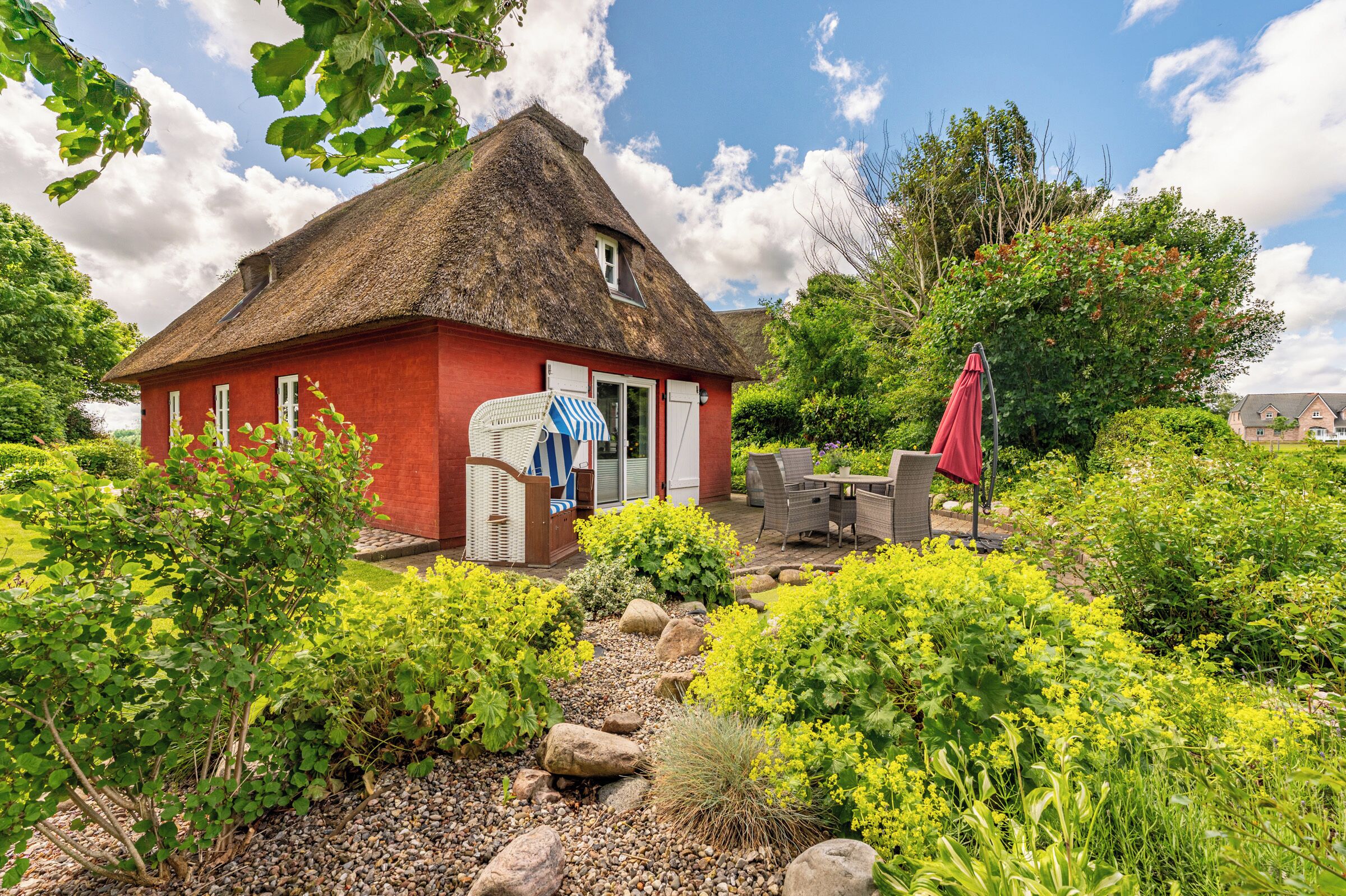 A red house with a thatched roof, a garden with various plants, and outdoor furniture.