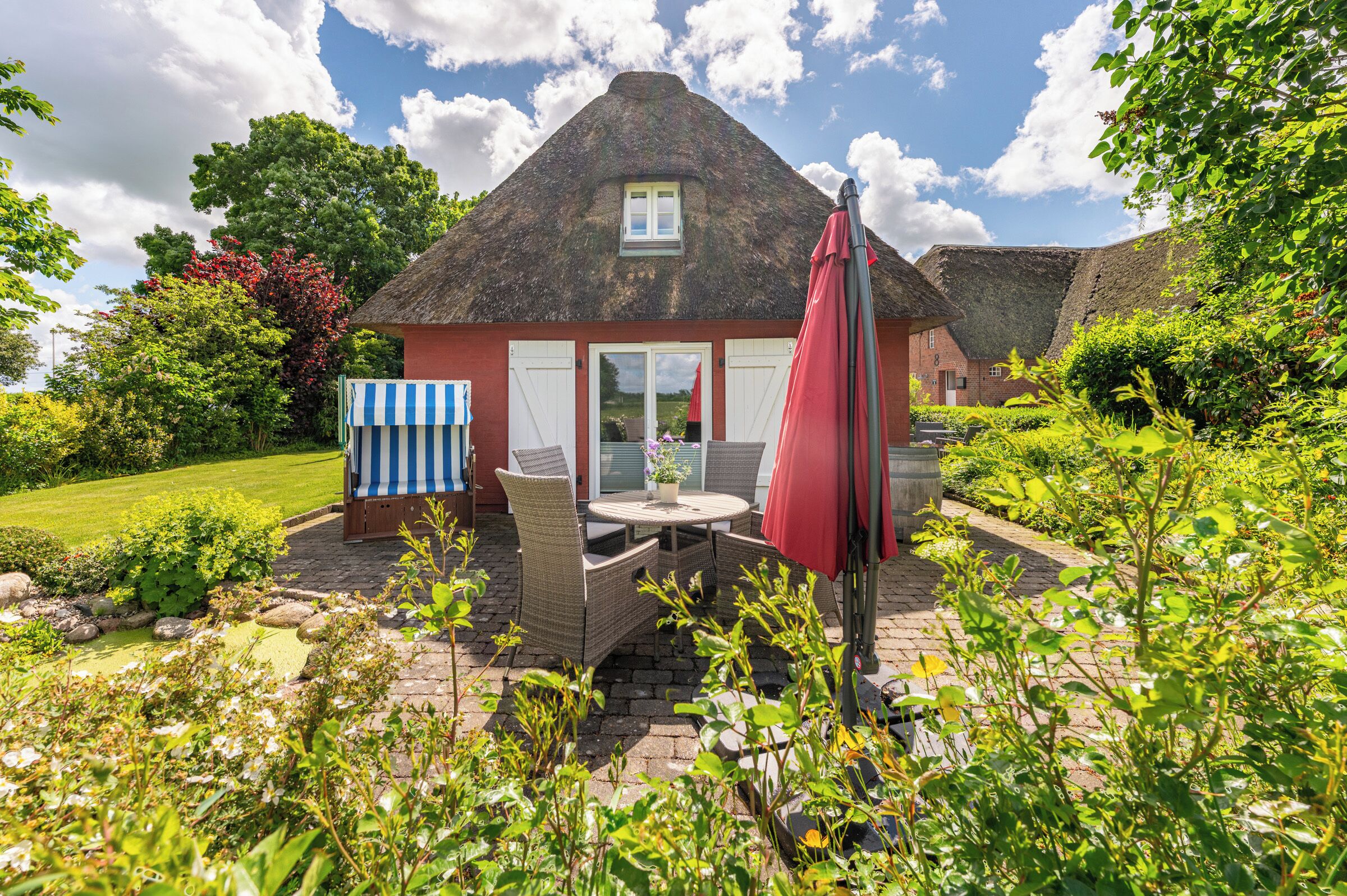 A thatched-roof house with a garden patio, a table with chairs, and a red umbrella.