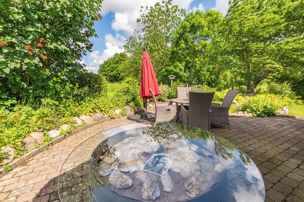 Une table ronde en verre, ornée de pierres et d’une photographie, est entourée d’une terrasse en briques, de meubles en osier et d’un parasol rouge.