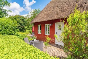 Une maison rouge avec un toit de chaume, une terrasse dotée de meubles d’extérieur et une végétation luxuriante qui entoure la propriété.