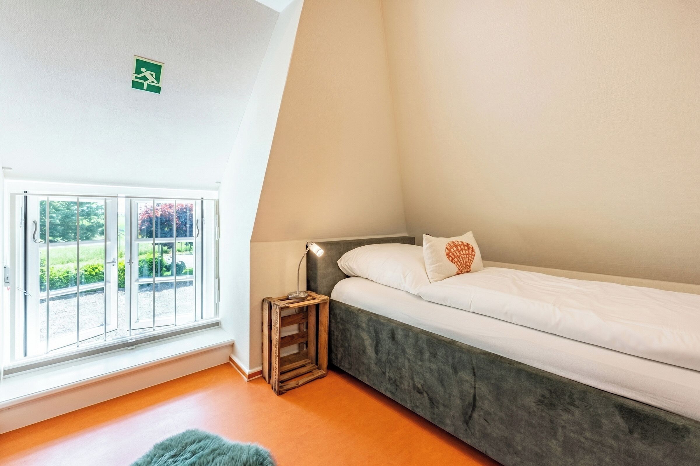 A bedroom with a bed, a window with a view of greenery, a small wooden side table, and a ceiling-mounted emergency exit sign.