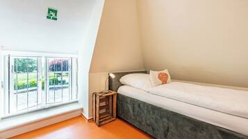 A bedroom with a bed, a window with a view of greenery, a small wooden side table, and a ceiling-mounted emergency exit sign.