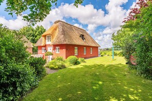 Une maison en briques rouges, coiffée d’un toit de chaume, entourée d’une pelouse bien entretenue et d’une végétation luxuriante.