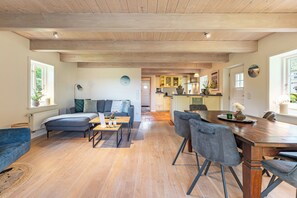 Un vaste espace de vie doté d’un plafond en bois, d’un canapé, d’une table à manger avec des chaises, et d’une cuisine à l’arrière-plan.