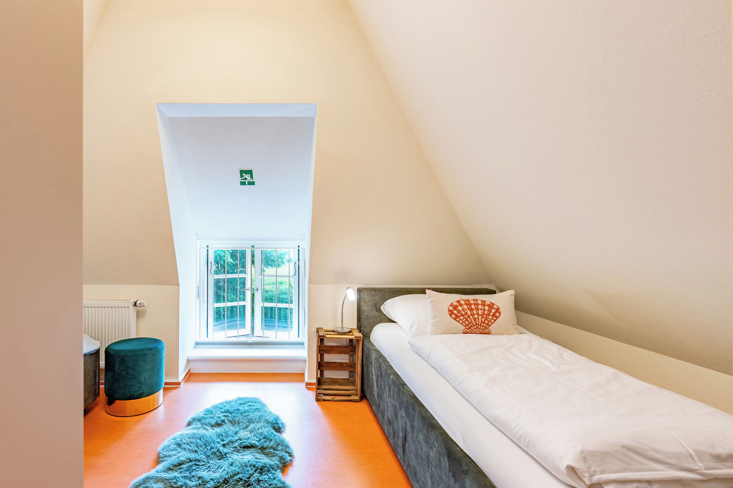A bedroom with a bed, a window, a radiator, a small stool, and a fluffy rug.