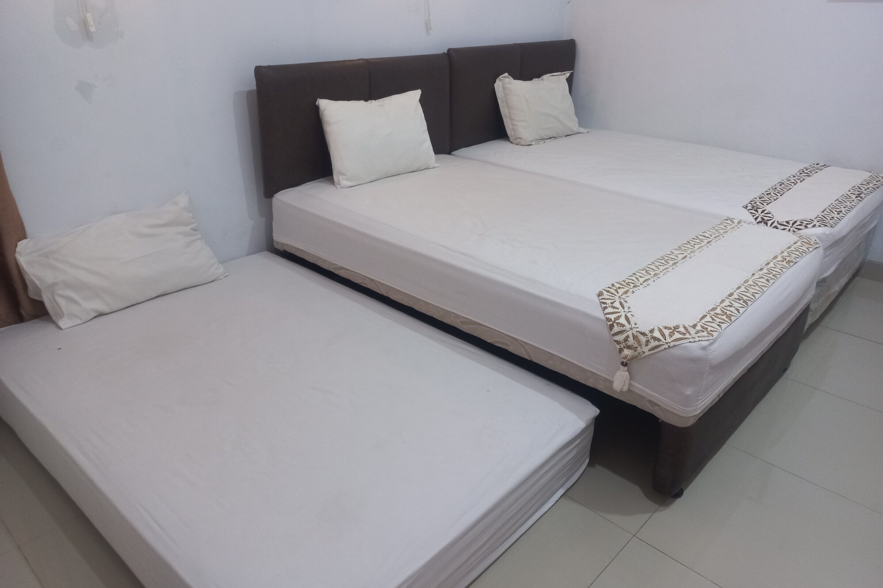 Un lit double avec une couvre-lit à motifs et deux oreillers, un lit simple plus petit avec une couvre-lit unie et un oreiller, et une tête de lit sombre.