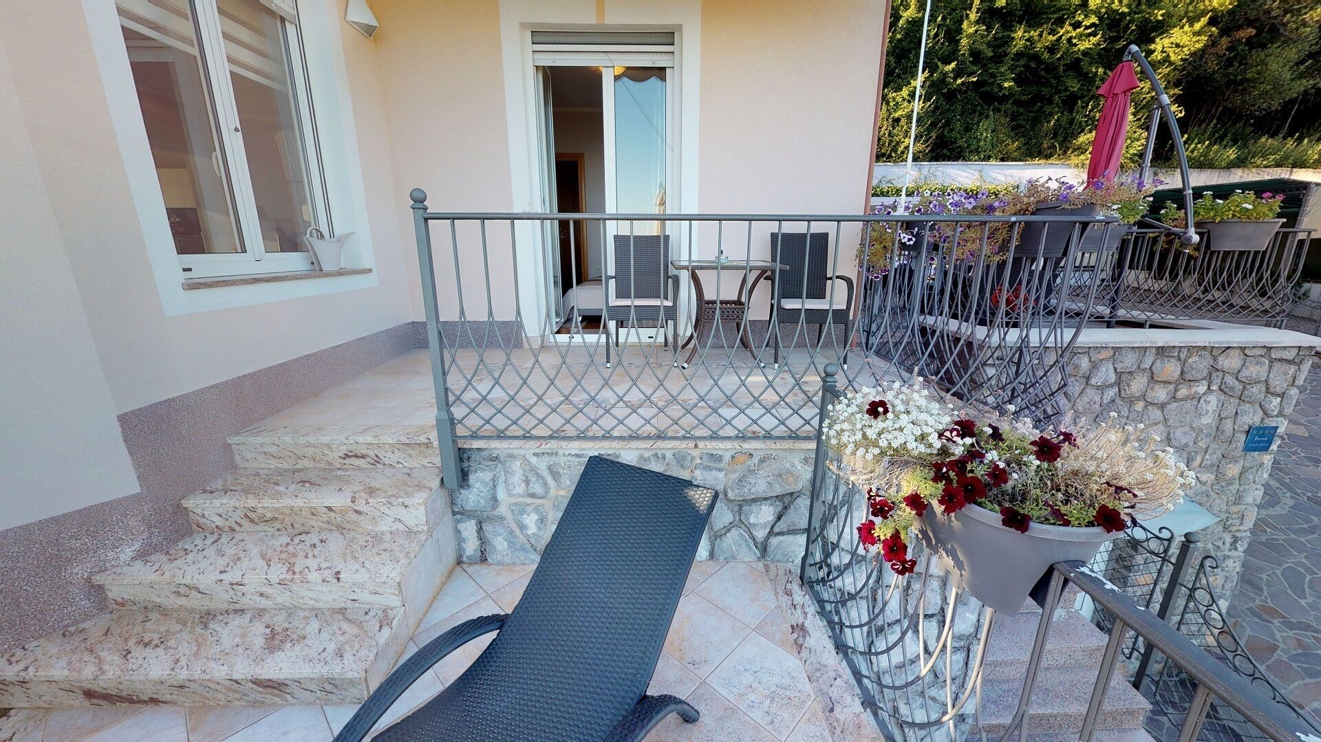 A balcony with a metal railing, a wicker chair, a table, and a potted plant with flowers.